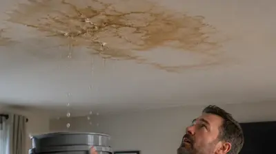Homeowner holding a bucket under a leaking ceiling with visible water stain after a storm, illustrating interior roof leak damage in a residential living room